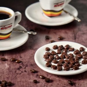 Chocolate covered espresso beans on a white dish with the presence of scattered espresso beans and two espresso cups of coffee.