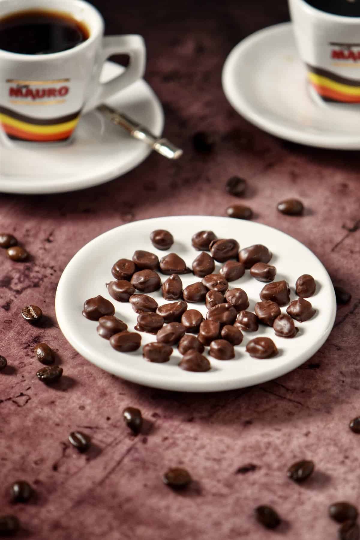 Chocolate covered espresso beans on a white dish with the presence of scattered espresso beans and two espresso cups of coffee.