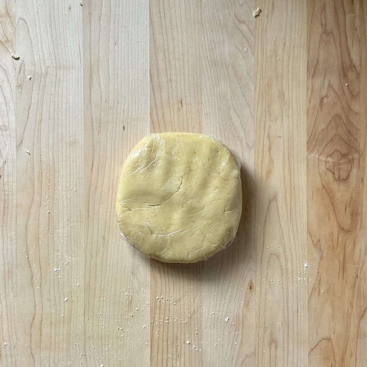 A disc of pasta frolla dough wrapped in cling wrap on a wooden board. 