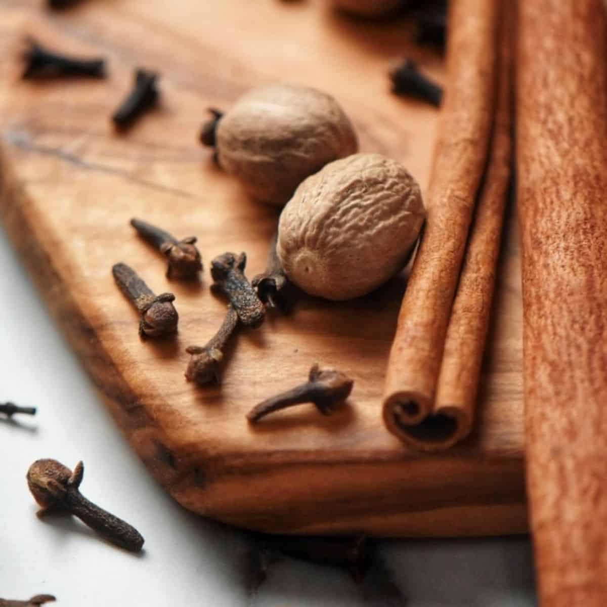 Long cinnamon stick on a wooden board, surrounded by whole nutmeg and cloves; the 3 ingredients required to make an allspice substitute.