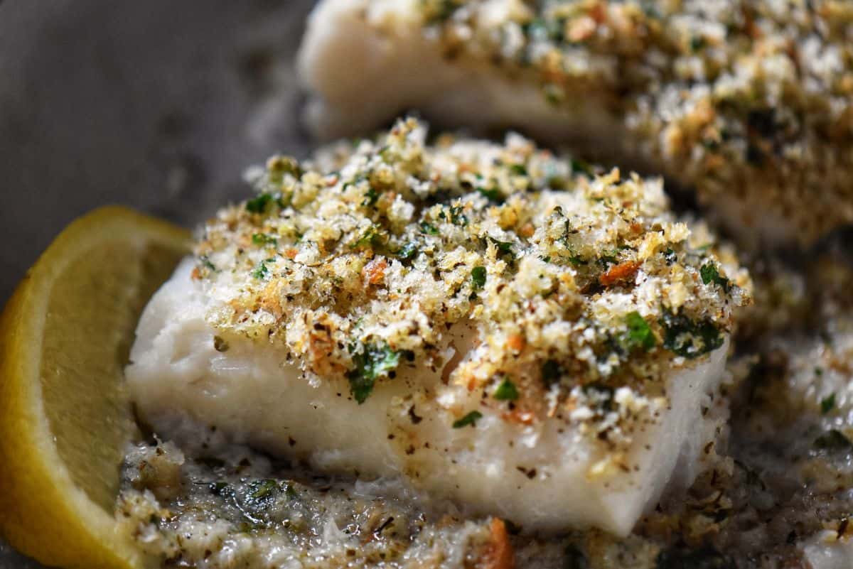 A close up of the baked cod fish on a white plate. Cod fish with seasoned breadcrumbs in a parchment lined pan.