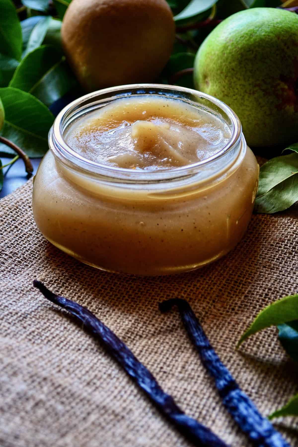 The Vanilla Pear Butter is seen in a wide open canning jar, surrounded by fresh pears.