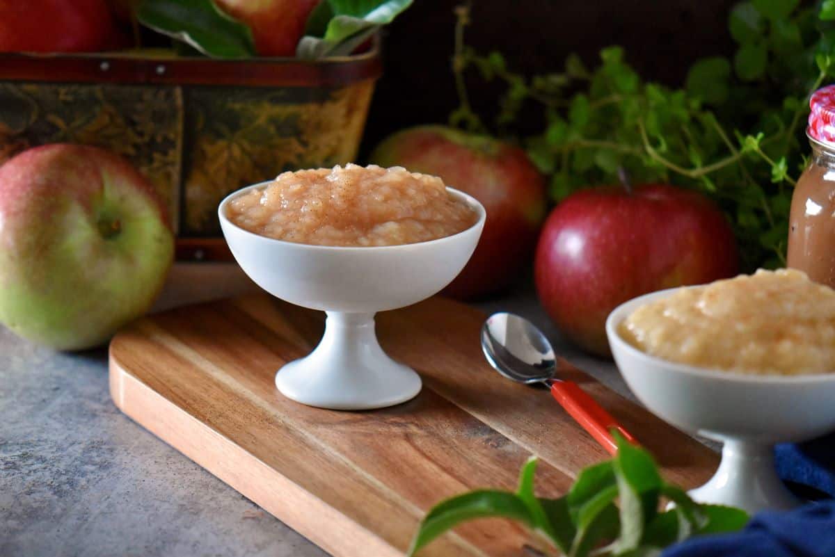 Crockpot applesauce in a white dessert dish, next to a spoon. In the background, a basket of apples.