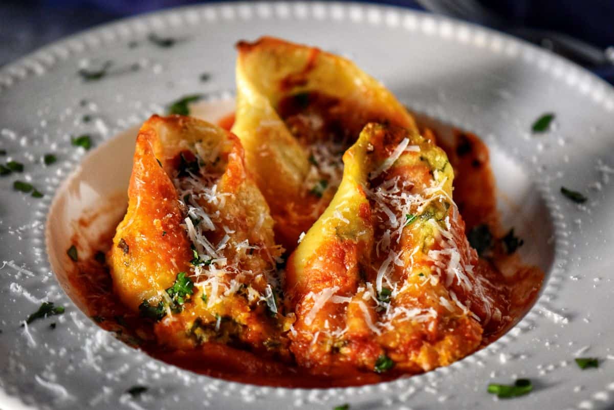 A close up photo of three giant ricotta cheese stuffed pasta shells on a white dinner plate garnished with grated cheese and chopped Italian parsley.