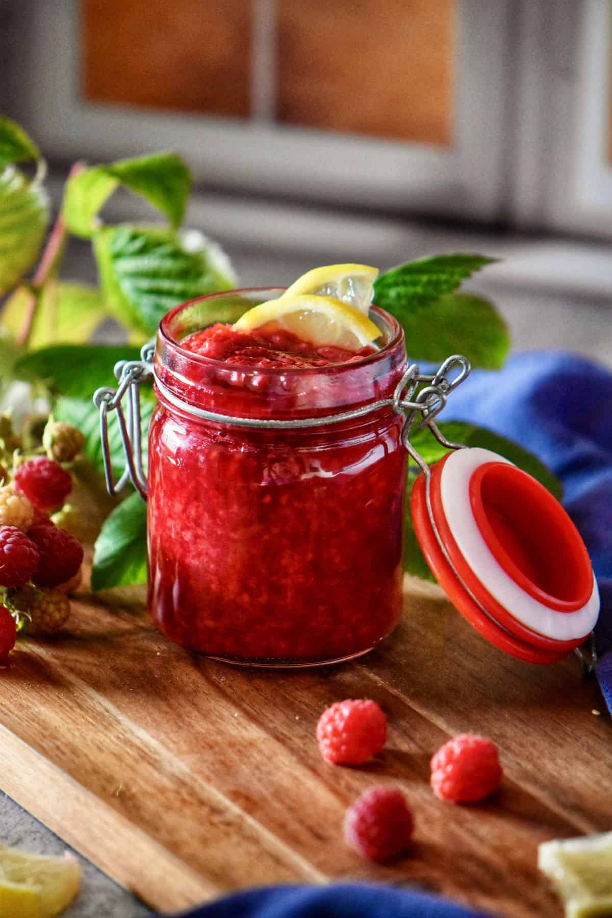 Homemade raspberry compote in a mason jar surrounded by fresh raspberries.
