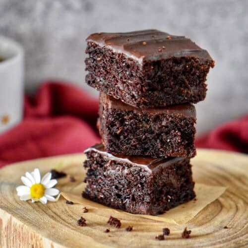 Squares of vegan chocolate zucchini brownies stacked on a wooden board, showing their fudgy texture.