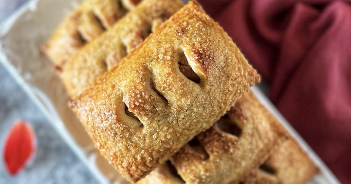 Golden brown apple hand pies with pasta frolla crust stacked on white plate, topped with cinnamon sugar.