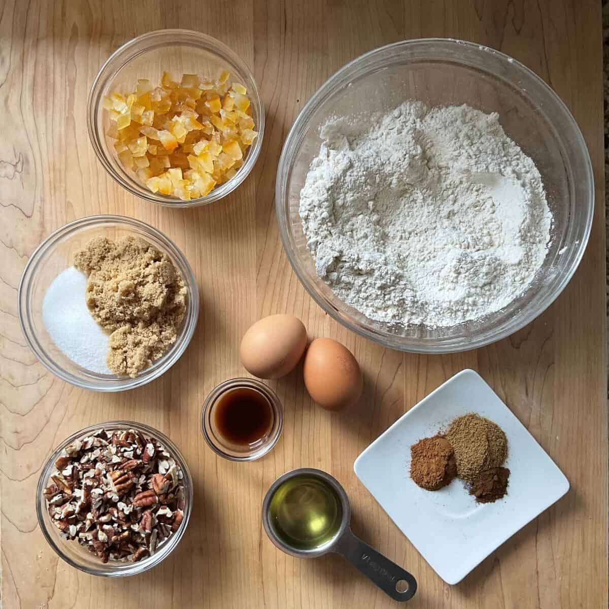 Ingredients for orange anise biscotti including flour, spices, candied orange peel, and toasted pecans arranged on a wooden surface.