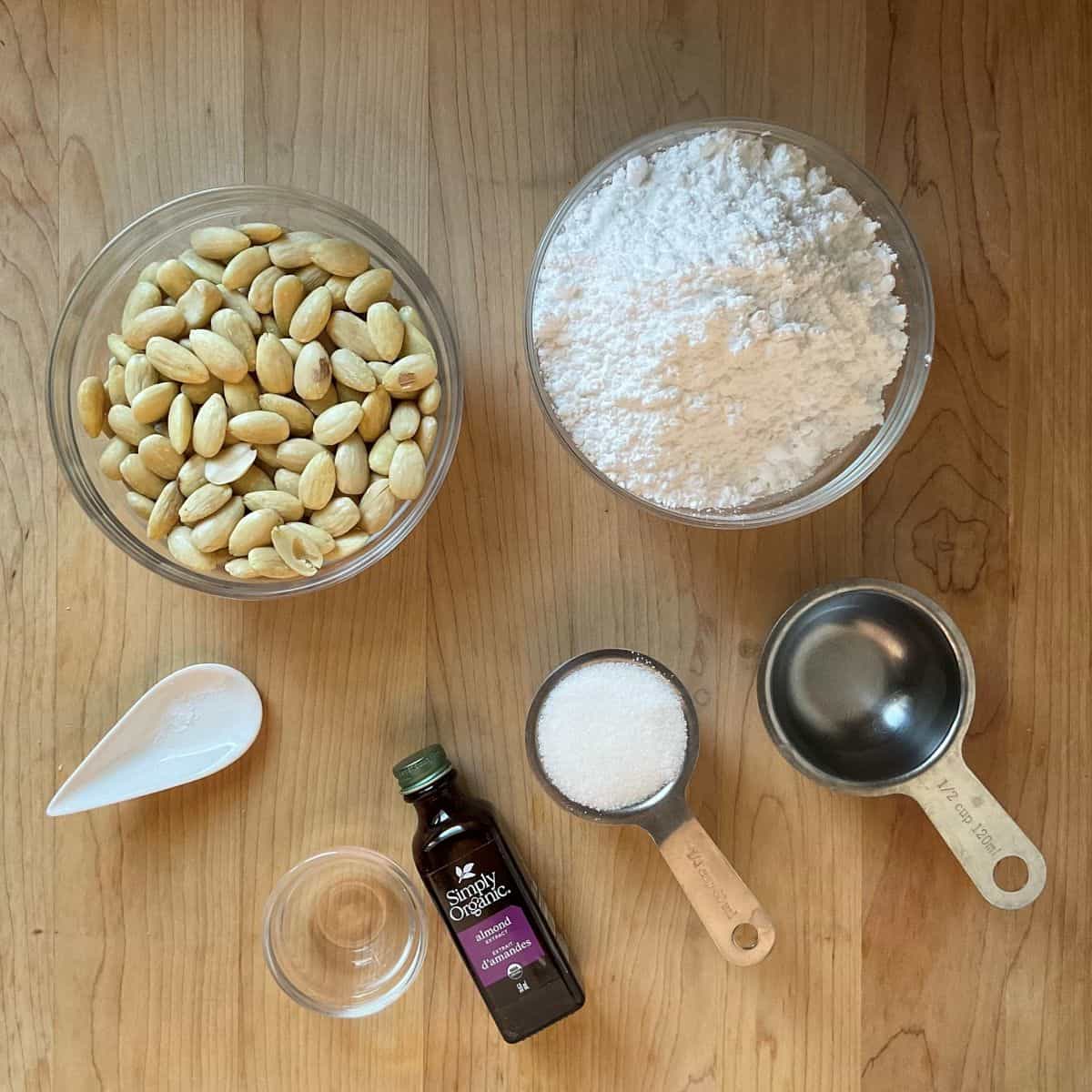Ingredients for homemade almond paste laid out on a countertop, including blanched almonds, powdered sugar, granulated sugar, water, and almond extract.