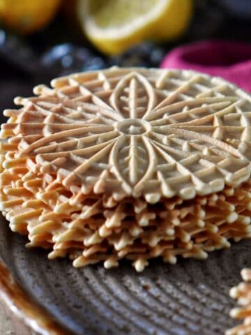 A stack of crisp lemon pizzelle cookies with fresh lemons in the background.