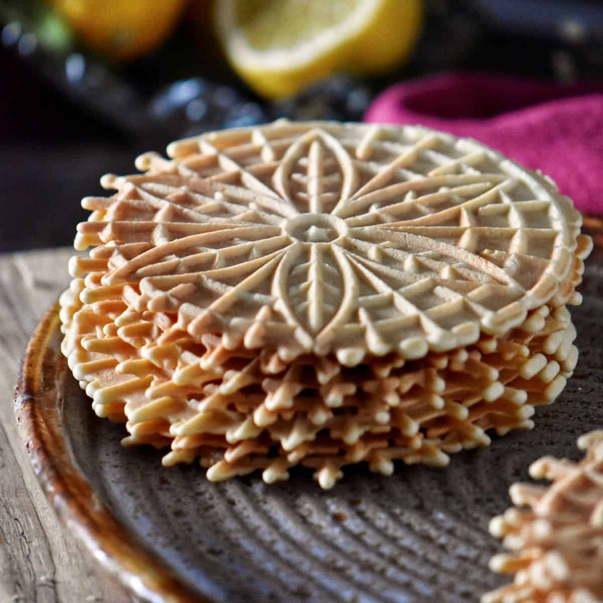 A stack of crisp lemon pizzelle cookies with fresh lemons in the background.
